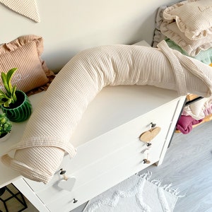 May include: A cream-colored waffle-textured pregnancy pillow in a C-shape, resting on a white dresser with three drawers. The pillow is on a white surface with a decorative rug in the foreground.