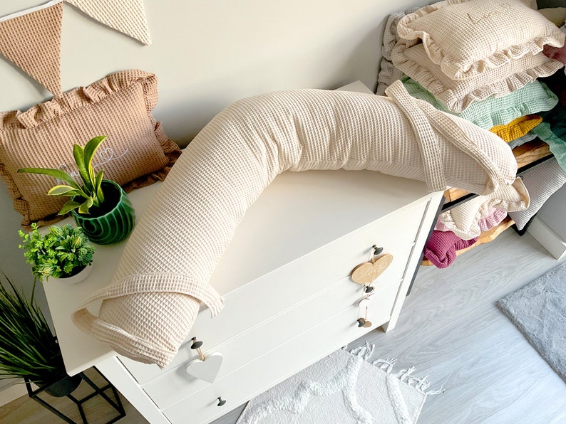 May include: A cream-colored waffle-textured pregnancy pillow in a C-shape, resting on a white dresser with three drawers. The pillow is on a white surface with a decorative rug in the foreground.