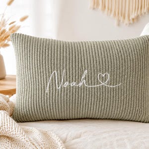 May include: A rectangular sage green knit cushion featuring a ribbed texture. The front is embroidered with the name Noah in white cursive script, followed by a small white heart outline. The cushion has a soft, woven fabric construction.