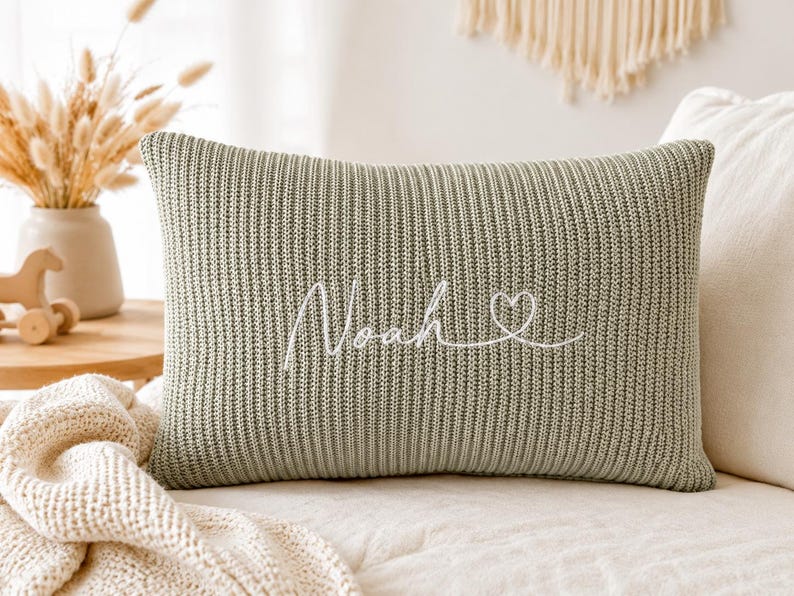May include: A rectangular sage green knit cushion featuring a ribbed texture. The front is embroidered with the name Noah in white cursive script, followed by a small white heart outline. The cushion has a soft, woven fabric construction.