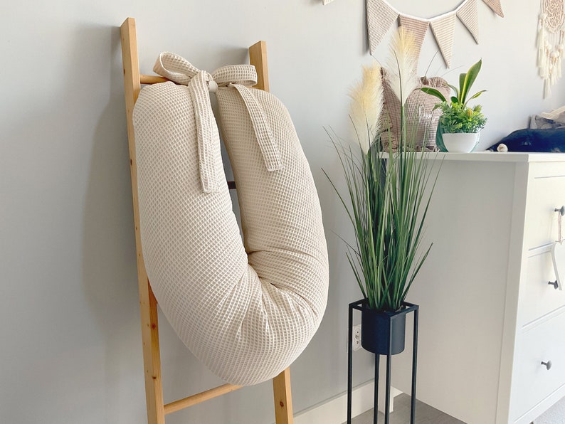 May include: A cream-colored waffle-textured pregnancy pillow shaped like a U, with a bow at the top, leaning against a wooden ladder.