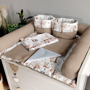 Peut inclure: Un matelas à langer blanc avec une bordure en tissu gaufré brun et un rouleau de matelas à langer en tissu gaufré brun. Le matelas à langer est imprimé d'animaux de la forêt blancs et bruns avec un tissu à motifs bruns, blancs et gris sur le dessus. Deux paniers en tissu gaufré brun avec un imprimé d'animaux de la forêt blancs et bruns sont sur le matelas à langer.