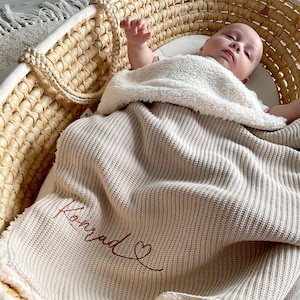 May include: A baby lies in a woven Moses basket, wrapped in a beige knitted blanket. The blanket is embroidered with the name "Konrad" in a cursive script, and a heart. A fluffy white blanket provides extra warmth.