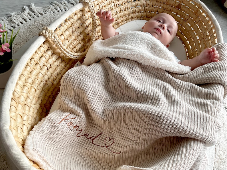 May include: A baby lies in a woven Moses basket, wrapped in a beige knitted blanket. The blanket is embroidered with the name "Konrad" in a cursive script, and a heart. A fluffy white blanket provides extra warmth.