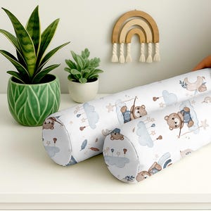 Könnte beinhalten: Zwei weiße zylindrische Babykissen mit einem sich wiederholenden Muster aus Teddybären, Wolken und Sternen. Die Kissen liegen auf einer weißen Oberfläche, mit grünen Topfpflanzen und einer Holz-Regenbogen-Dekoration im Hintergrund.