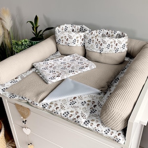 Op de afbeelding: Een beige verschoningsset met een bloemenprint. De set bevat een verschoningsmat, twee bolsterkussens, een klein dekentje en twee opbergmanden. De verschoningsmat is gemaakt van een wafelstof en voelt zacht en pluche aan.