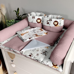 Peut inclure: Une table à langer pour bébé avec un matelas à langer rose gaufré, des coussins de protection et deux paniers de rangement. Les paniers et une petite couverture présentent un imprimé animalier de safari. Une table à langer blanche est visible.