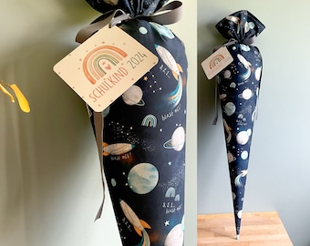 Può includere: Un cono scolastico nero e blu a tema spaziale. Il cono è decorato con pianeti, stelle, razzi e il testo "Blast off!" e "3, 2, 1... Blast off!" Un'etichetta attaccata al cono recita "Schulkind 2024" con una grafica arcobaleno.