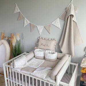 Peut inclure: Un lit bébé blanc avec un ensemble de literie beige et blanc. La literie comprend un oreiller brodé du nom "Paul", un pare-chocs et un drap-housse. Il y a une guirlande beige et blanche suspendue au-dessus du lit.