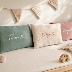 Peut inclure: Trois coussins rectangulaires en tissu mousseline texturé, de couleurs vert sauge, crème et vieux rose. Chaque coussin est brodé avec les prénoms Fiona, Elizan et Zoey, accompagnés d'une petite icône en forme de cœur sur le côté droit.