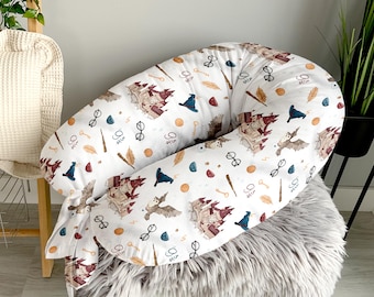 May include: A white, U-shaped nursing pillow with a repeating pattern of brown castles, owls, eyeglasses, keys, and feathers. The pillow is designed for comfort and support during feeding and is placed on a gray, fluffy surface.