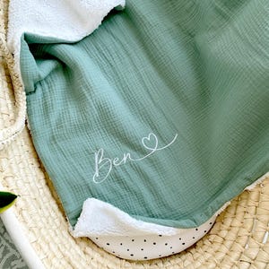 May include: A soft, green muslin baby blanket with a white sherpa lining. The blanket has the name "Ben" embroidered on it with a small heart.