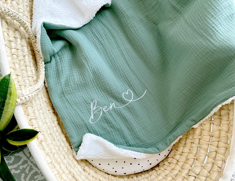May include: A soft, green muslin baby blanket with a white sherpa lining. The blanket has the name "Ben" embroidered on it with a small heart.