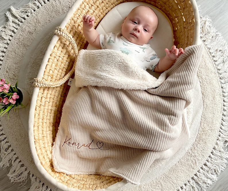 May include: A baby lies in a woven bassinet, covered by a beige blanket embroidered with the name "Konrad". The blanket has a soft, plush lining. The bassinet rests on a round rug with a fringed edge.