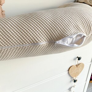 May include: A white waffle-textured cylindrical pillow with a zipper opening. The pillow is lying on a white dresser with a wooden heart hanging from a knob.