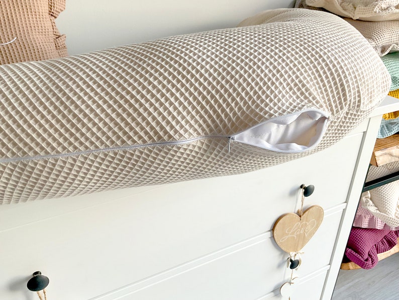 May include: A white waffle-textured cylindrical pillow with a zipper opening. The pillow is lying on a white dresser with a wooden heart hanging from a knob.