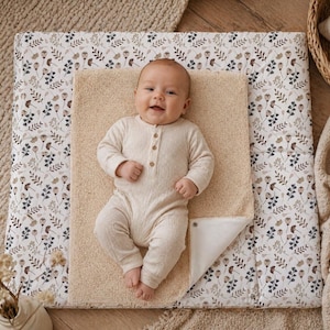 Könnte beinhalten: Ein lächelndes Baby in einem cremefarbenen Strampler liegt auf einer beigen Wickelunterlage. Die Unterlage befindet sich auf einer größeren Wickelmatte mit floralem Muster in Weiß, Braun und Blau. Ein Weidenkorb und eine Decke sind im Hintergrund.