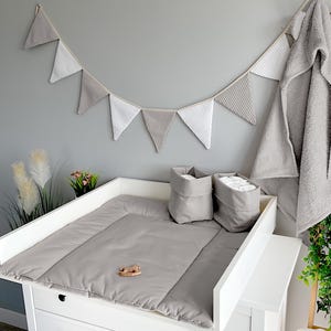 Könnte beinhalten: Ein weißer Wickeltisch mit einer grauen Wickelauflage, zwei grauen Aufbewahrungskörben und einem Holzspielzeug. Über dem Tisch hängt eine graue Girlande, und rechts befindet sich ein graues Kapuzenhandtuch.