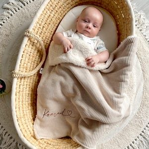May include: A baby lies in a woven bassinet, covered with a beige blanket. The blanket has a soft, plush lining and is embroidered with the name "Konrad" in cursive. The bassinet sits on a round, cream-coloured rug.