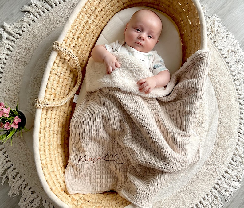 May include: A baby lies in a woven bassinet, covered with a beige blanket. The blanket has a soft, plush lining and is embroidered with the name "Konrad" in cursive. The bassinet sits on a round, cream-coloured rug.