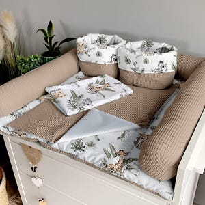 Peut inclure: Un ensemble de matelas à langer pour bébé avec un imprimé animalier safari. L'ensemble comprend un matelas à langer, deux coussins de soutien et deux paniers de rangement. Le matelas à langer est bleu clair avec un fond blanc et présente une variété d'animaux de la savane, notamment des girafes, des lions et des zèbres. Les coussins de soutien sont beiges et ont une texture gaufré. Les paniers de rangement sont blancs avec une bordure beige et présentent un imprimé animalier safari.