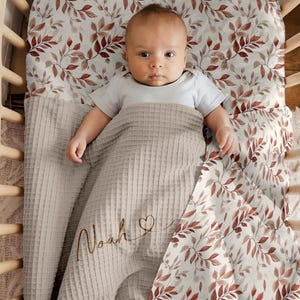 May include: A baby is lying in a cot with a grey waffle weave blanket and a white and red floral sheet. The blanket has the name "Noah" embroidered on it.