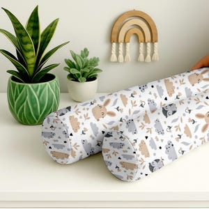 Puede incluir: Dos protectores cilíndricos para bebés con fondo blanco y un estampado repetido de conejitos, nubes y hojas en tonos beige, gris y negro. Los protectores están sobre una superficie blanca, con una planta verde en maceta y un arcoíris de madera en el fondo.