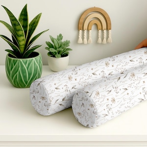 Op de afbeelding: Twee witte cilindrische kussens met een bloemmotief in bruin en beige. De kussens liggen op een wit oppervlak. Een groene plant in een pot en een houten regenboogdecoratie zijn op de achtergrond.
