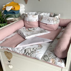 Könnte beinhalten: Eine weiße Wickelauflage mit einem rosa-weißen Elefantenmuster. Die Auflage ist umgeben von rosa, wattierten Stoßfängern und zwei weißen Körben mit rosa, wattierten Deckeln. Die Körbe haben ein rosa-weißes Elefantenmuster.