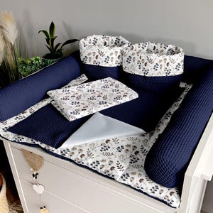 Peut inclure: Un ensemble de matelas à langer bleu marine avec un imprimé floral blanc. L'ensemble comprend un matelas à langer, deux coussins de soutien, une petite couverture et deux paniers de rangement.
