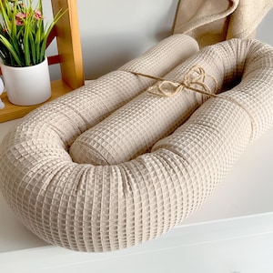 Op de afbeelding: Een beige baby nestje met wafeltextuur, vastgebonden met touw. Het nestje is U-vormig, ontworpen voor babycomfort. De achtergrond bevat een houten plank met een plant en een decoratief konijn nachtlampje.