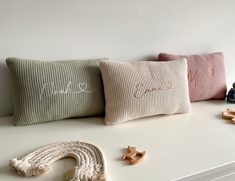 May include: Three rectangular knit cushions in sage green, cream, and dusty rose. Each cushion features embroidered cursive names: Noah, Emma, and a third name, accompanied by a small heart outline. The cushions have a ribbed texture.