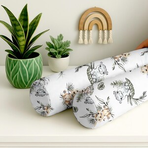 Puede incluir: Dos almohadas cilíndricas blancas con un patrón repetido de hipopótamos grises, flores color beige y hojas verdes. Las almohadas están sobre una superficie blanca, con plantas en macetas verdes y una decoración de arcoíris de madera en el fondo.