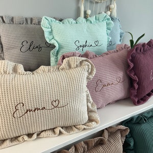 May include: A set of six decorative cushions with ruffled edges. The cushions are made from a waffle-textured fabric and feature embroidered names, including Elias, Sophia, Emma, Lina, and Mia. Each name is accompanied by a small heart.