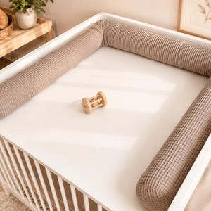 Op de afbeelding: Een wit aankleedkussen voor op de commode met een taupekleurige wafelstof rand die drie zijden omsluit. Een kleine, lichtgekleurde houten rammelaar met een centrale kraal ligt in het midden van het gladde, vlakke witte oppervlak.