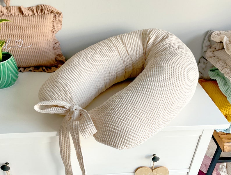 May include: A cream-colored waffle-textured pregnancy pillow with a long tie. The pillow is shaped like a donut and is resting on a white dresser.