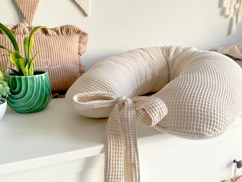 May include: A cream-colored waffle-textured pregnancy pillow with a long tie. The pillow is shaped like a horseshoe and is tied with a bow.