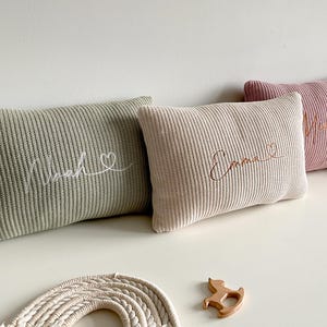 May include: Three rectangular knit cushions in sage green, cream, and dusty rose. Each cushion features embroidered cursive text and a small heart symbol. The first cushion reads Noah, the second reads Emma, and the third has partially visible text.