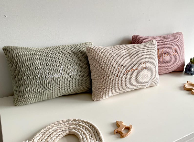 May include: Three rectangular knit cushions in sage green, cream, and dusty rose. Each cushion features embroidered cursive text and a small heart symbol. The first cushion reads Noah, the second reads Emma, and the third has partially visible text.