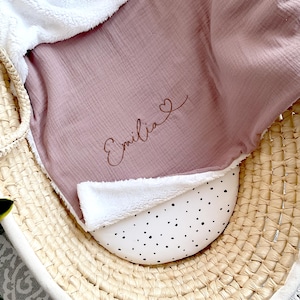 May include: A pink muslin baby blanket with a white sherpa trim. The blanket is personalised with the name "Emilia" and a small heart.