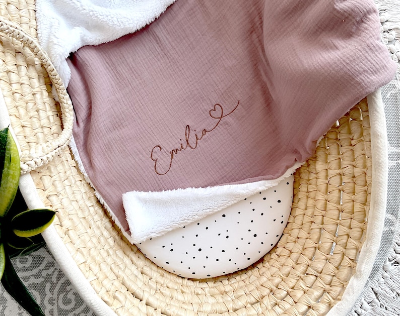 May include: A pink muslin baby blanket with a white sherpa trim. The blanket is personalised with the name "Emilia" and a small heart.