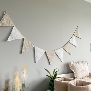 Könnte beinhalten: Ein beige-weißes Dreiecksbanner mit gewelltem Rand hängt an einer Wand. Das Banner ist aus Stoff und hat einen rustikalen, ländlichen Stil.