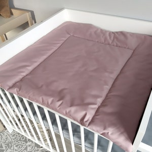 Könnte beinhalten: Ein weißes Babybett mit einer rosa gesteppten Wickelauflage oben. Die Wickelauflage hat eine weiche, plüschige Textur und ist so konzipiert, dass sie eine komfortable und sichere Oberfläche zum Wickeln eines Babys bietet.