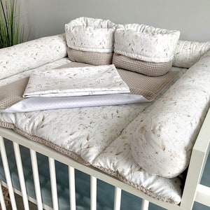 Könnte beinhalten: Ein weißes Babybett mit einer beige-weißen, floral gemusterten Matratze und Stoßstangen. Das Bett enthält außerdem eine beige Waffel-Decke und zwei beige-weiße, floral gemusterte Körbe.