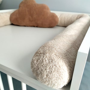 Può includere: Un lettino bianco con un paraurti in peluche beige e un cuscino a forma di nuvola marrone. Il paraurti è realizzato in un materiale morbido e soffice ed è progettato per proteggere il bambino da urti e cadute.