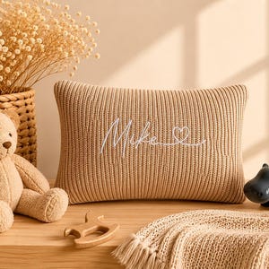 May include: A rectangular beige knit cushion featuring the embroidered white script name Mike followed by a small heart outline. The cushion is positioned next to a light brown teddy bear, a wooden toy, and a dark grey hippo-shaped planter.