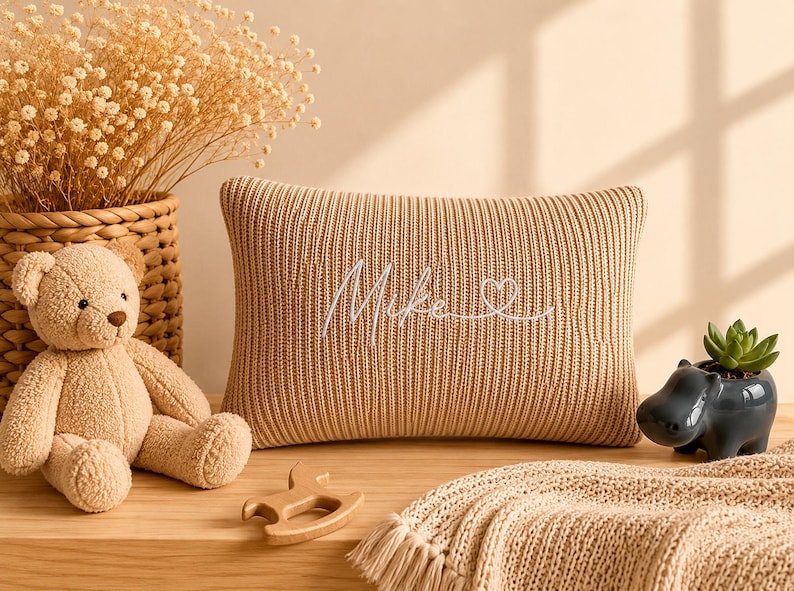 May include: A rectangular beige knit cushion featuring the embroidered white script name Mike followed by a small heart outline. The cushion is positioned next to a light brown teddy bear, a wooden toy, and a dark grey hippo-shaped planter.