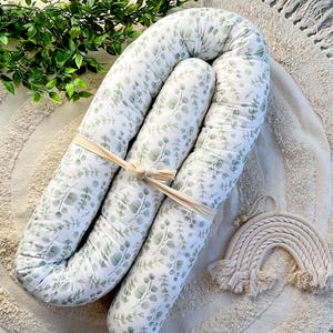 Peut inclure: Un nid de bébé blanc et vert avec un motif de feuilles d'eucalyptus, noué avec un ruban beige. Une décoration arc-en-ciel en macramé est placée à côté du nid. Le nid est sur un tapis crème.