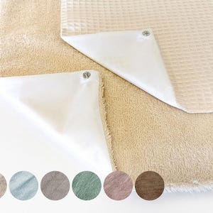 Może przedstawiać: Miękki, beżowo-biały koc z dwoma białymi kwadratami z tkaniny, przymocowanymi za pomocą zatrzasków. Kwadraty mają gładkie, satynowe wykończenie. Koc ma miękką, puszystą fakturę.