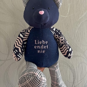 Puede incluir: Un oso de peluche confeccionado con varios tejidos estampados. La cabeza y el torso son de color azul marino oscuro, con el torso mostrando el texto bordado 'Liebe endet nie'. Las orejas, brazos y piernas presentan patrones geométricos y de cuadros en negro, blanco y rosa.
